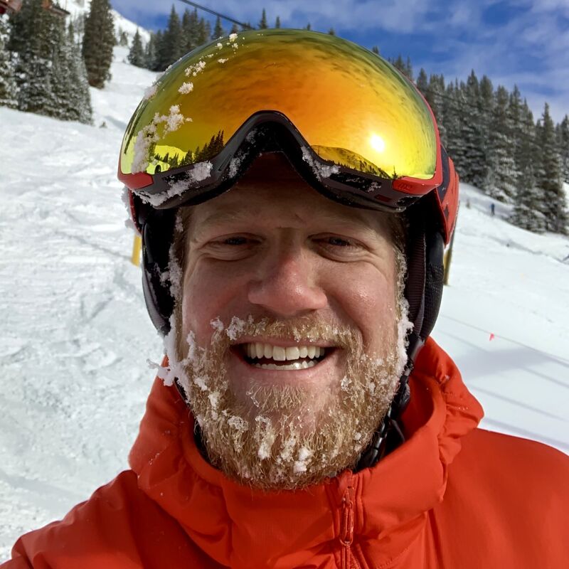 The image shows a man in ski gear, including a helmet and goggles, with snow or frost clinging to his beard. He is smiling and appears to be on a snowy mountain, likely skiing or snowboarding. The background features snow-covered trees and a clear sky, suggesting a cold but sunny day on the slopes. He is wearing an orange jacket.